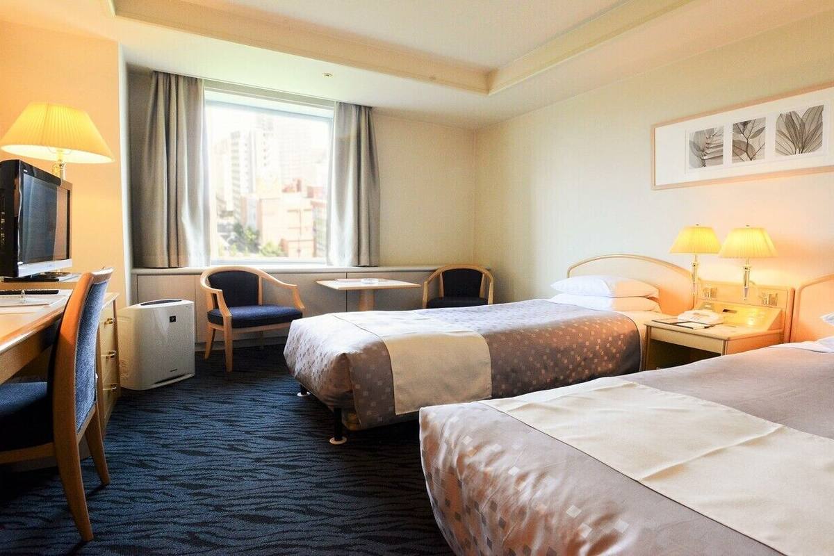 A hotel room with two beds, a desk with a computer, a chair, a small table, and a window with curtains.