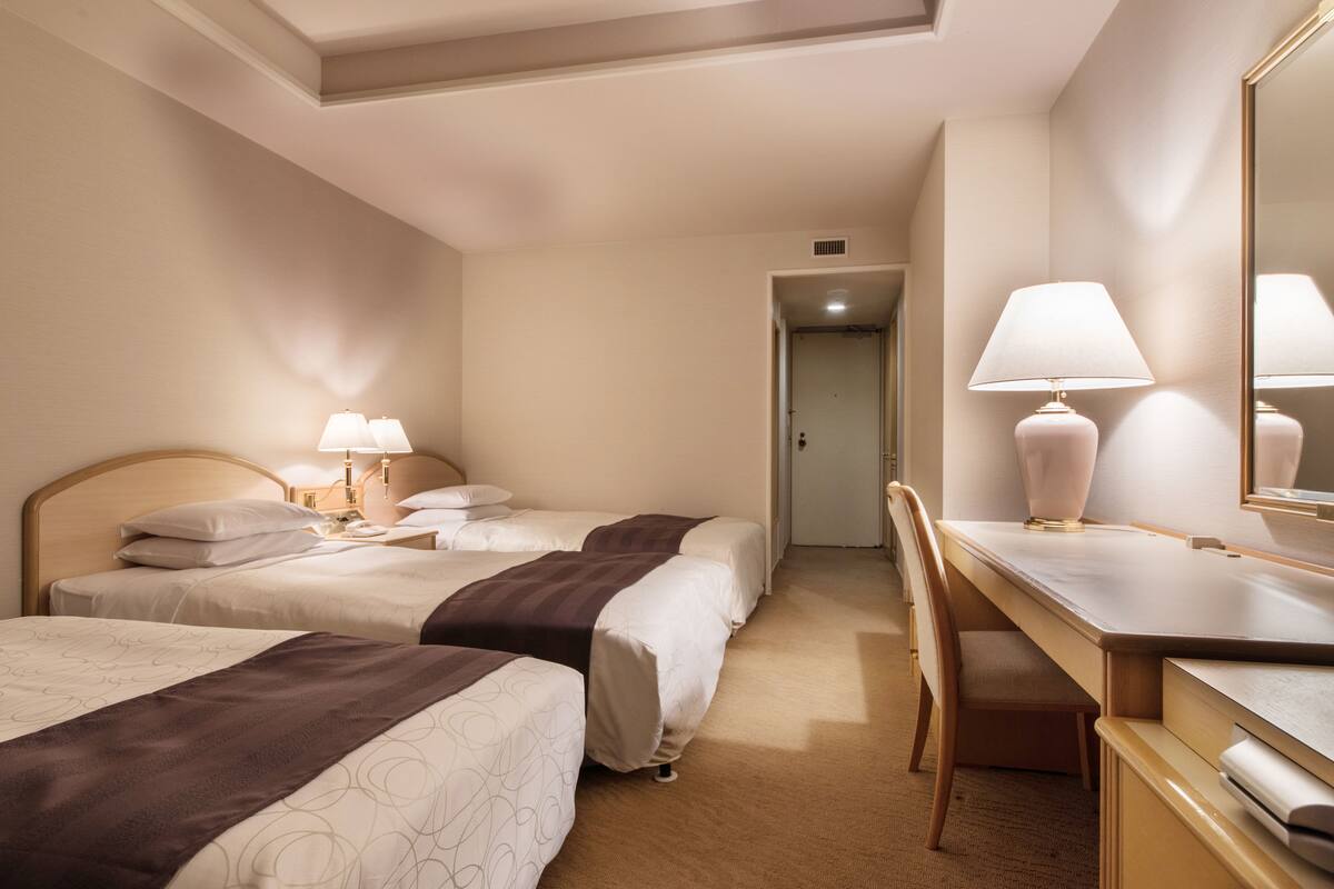 A hotel room with two beds, a desk, a chair, a lamp, and a mirror.