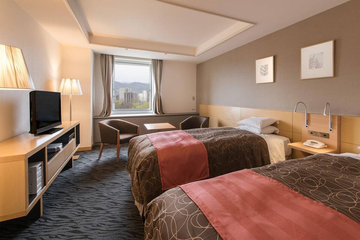 A hotel room with two beds, a desk, a TV, and a window with curtains.