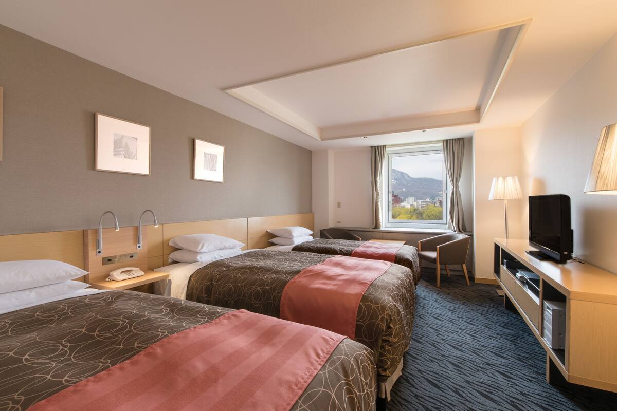 A hotel room with three beds, a desk, a television, and a window with a view of the city.