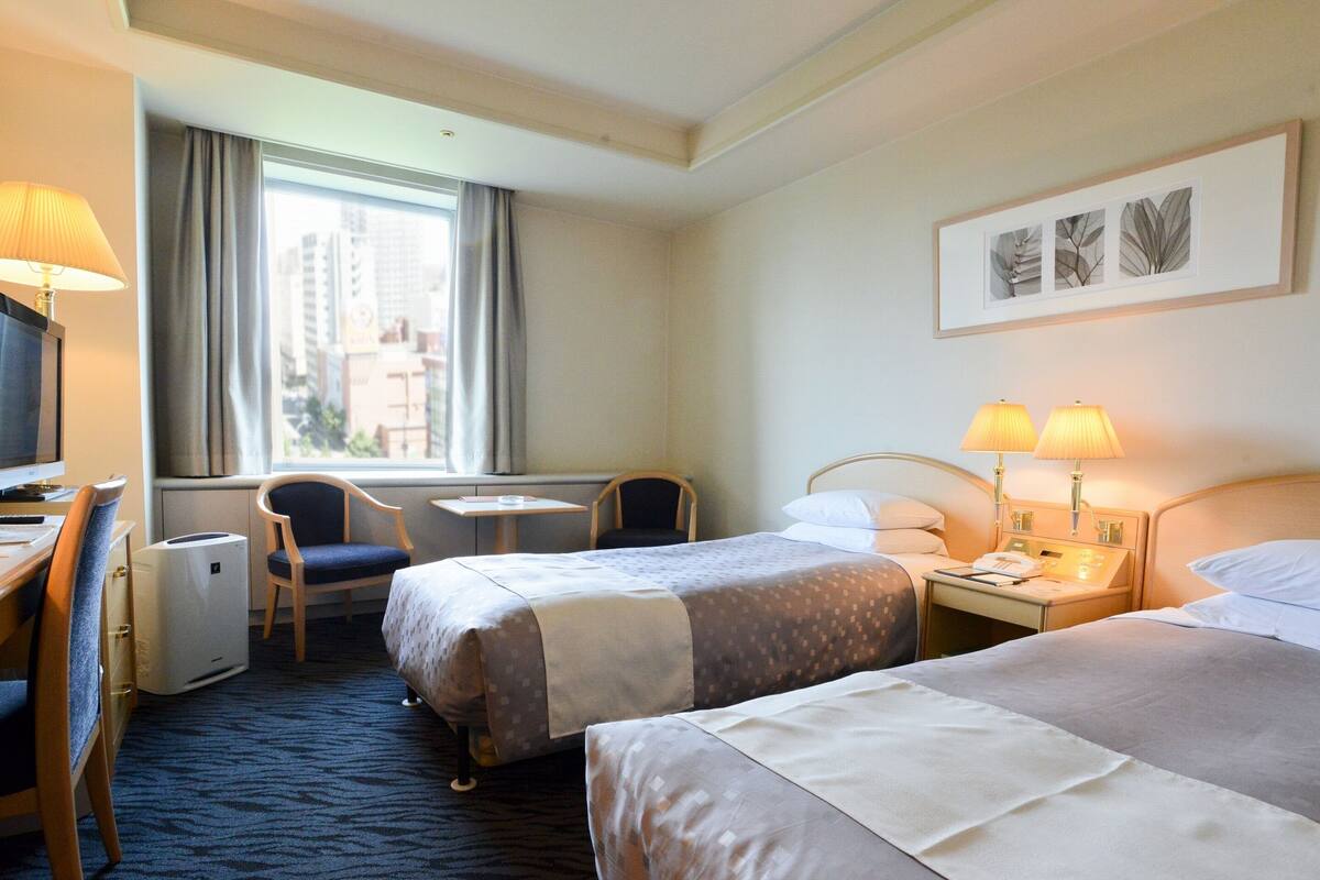 A hotel room with two beds, a desk with a computer, a chair, a small table, and a window with curtains.