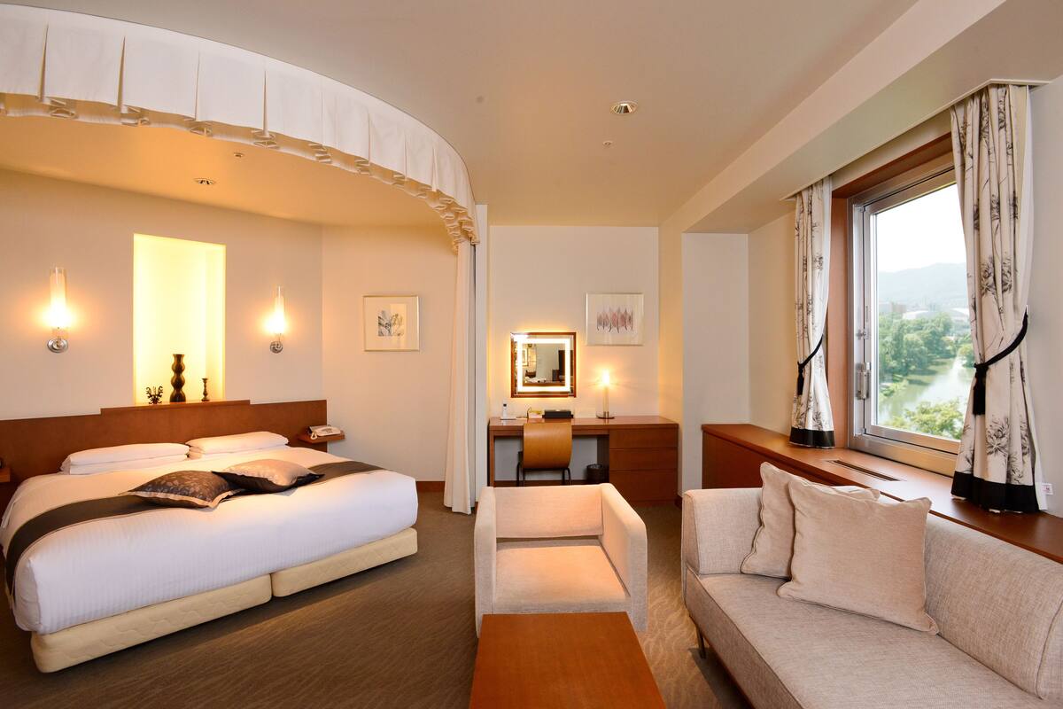 A hotel room with a large bed, a desk with a chair, a sofa, and a window with curtains.