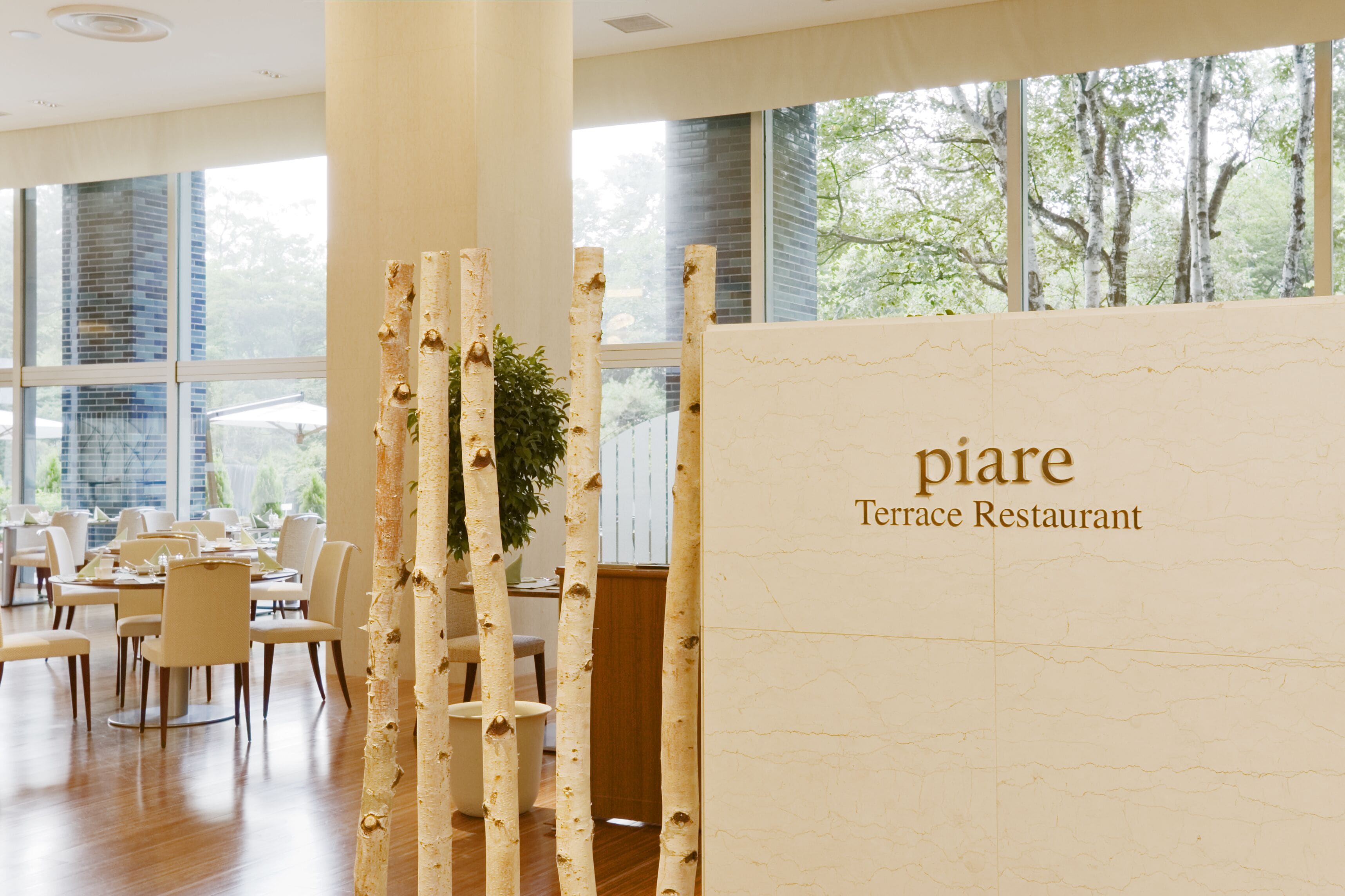 A restaurant interior with a modern design, featuring birch tree-like columns and a view of the outdoors through large windows.