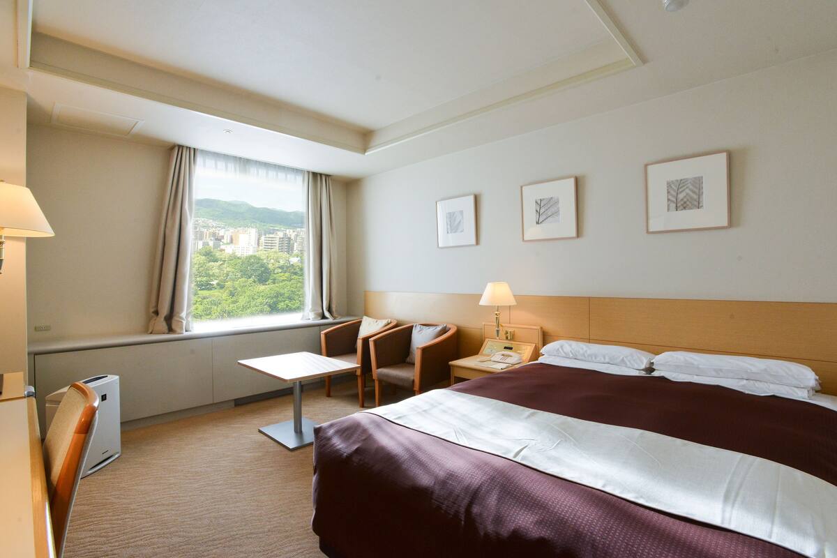 A hotel room with a large bed, a desk, a chair, and a window with a city view.