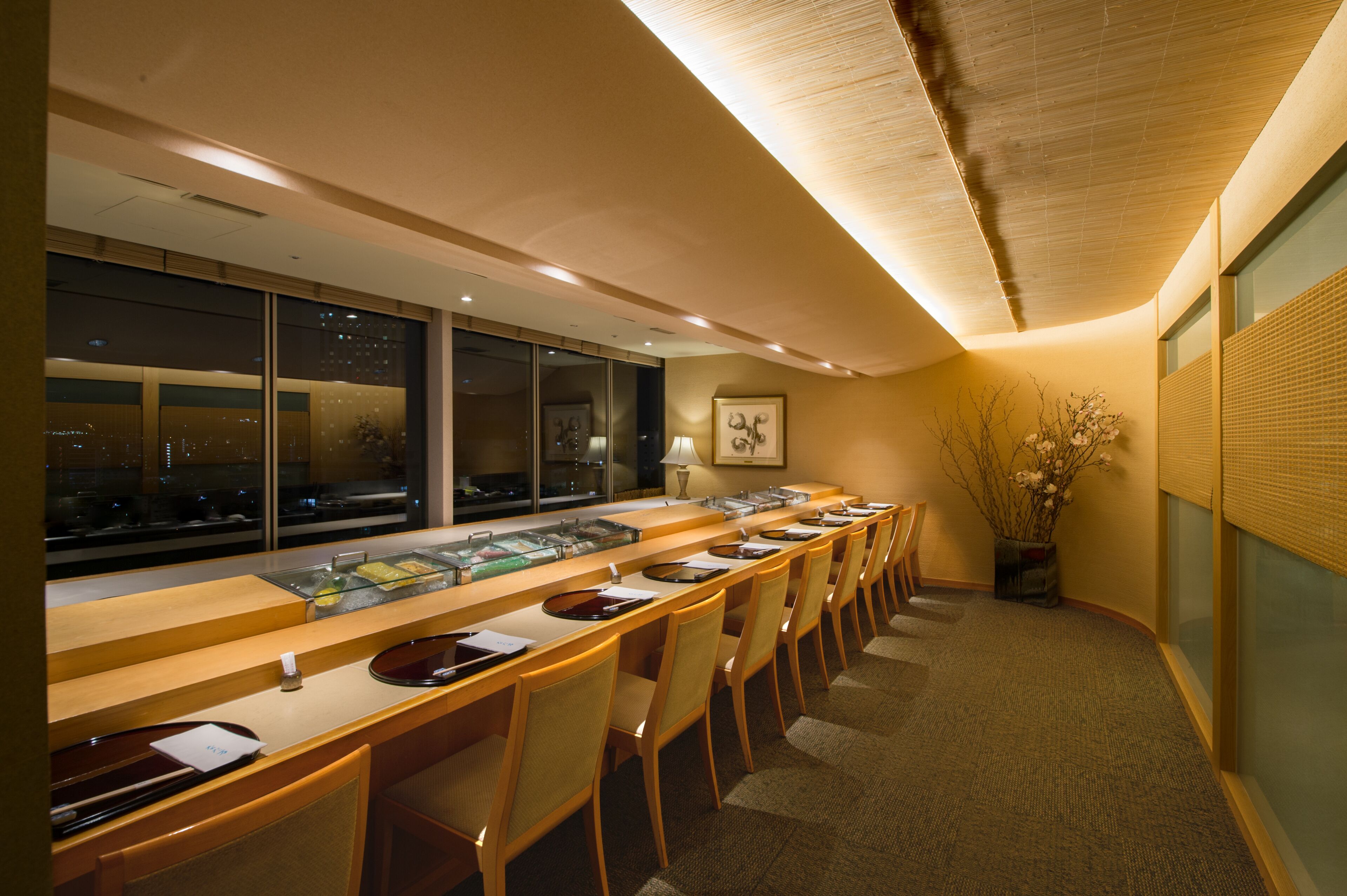 A hotel reception area with a long counter, multiple chairs, and a large window offering a view of the cityscape at night.