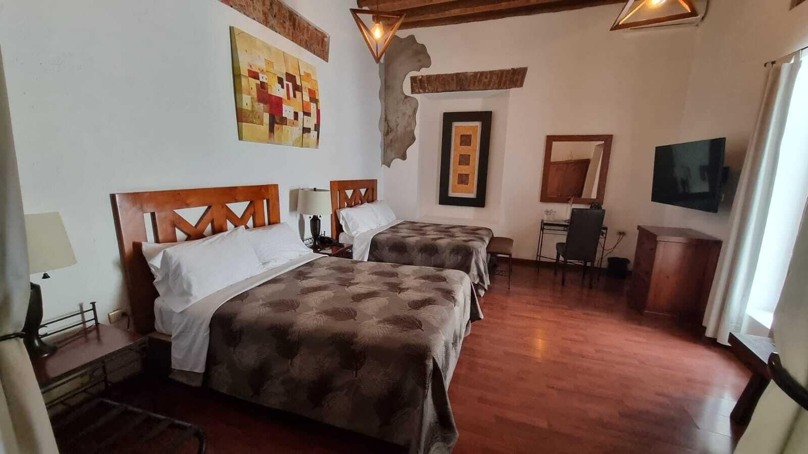 A hotel room with two beds, a desk, a chair, a TV, and artwork on the walls.