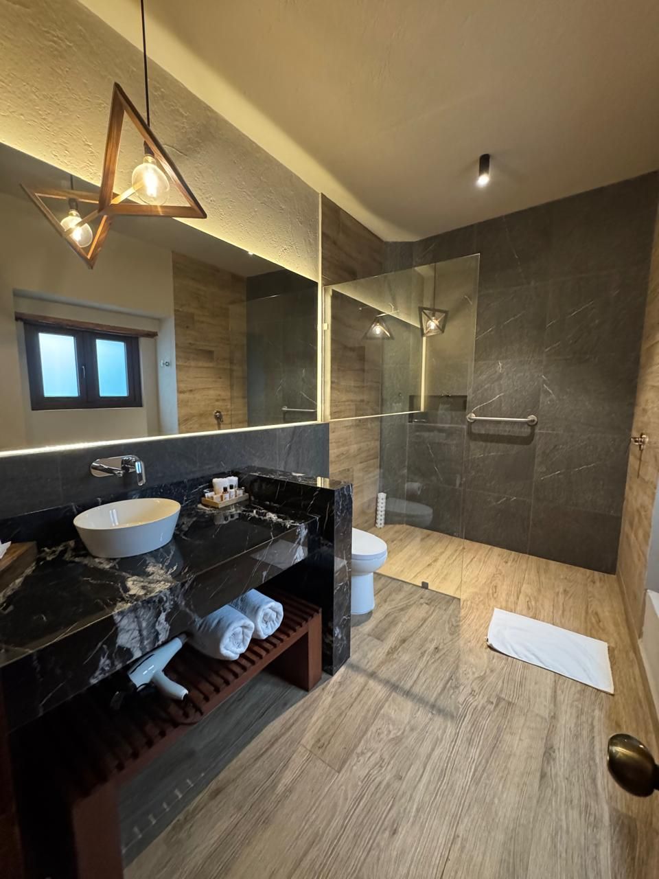 A modern bathroom with a large mirror, a round sink, a shower area, and a wooden floor.