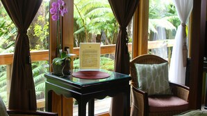 A room with a wooden table, a wicker chair, and a view of lush greenery through large windows.