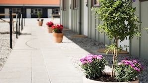Un chemin menant à un bâtiment doté d’un toit rouge, de plantes en pot ornées de fleurs roses et d’un petit arbre.