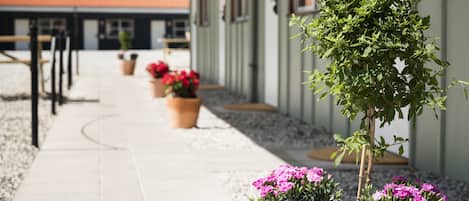 A pathway leading to a building with a red roof, potted plants with pink flowers, and a small tree.