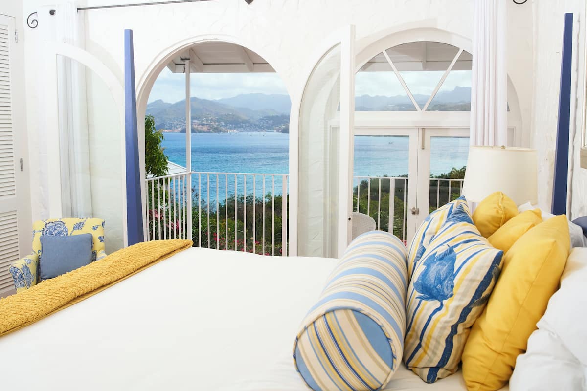 A bedroom with a bed, yellow and blue striped pillows, a yellow throw blanket, and a view of the sea through an arched window.