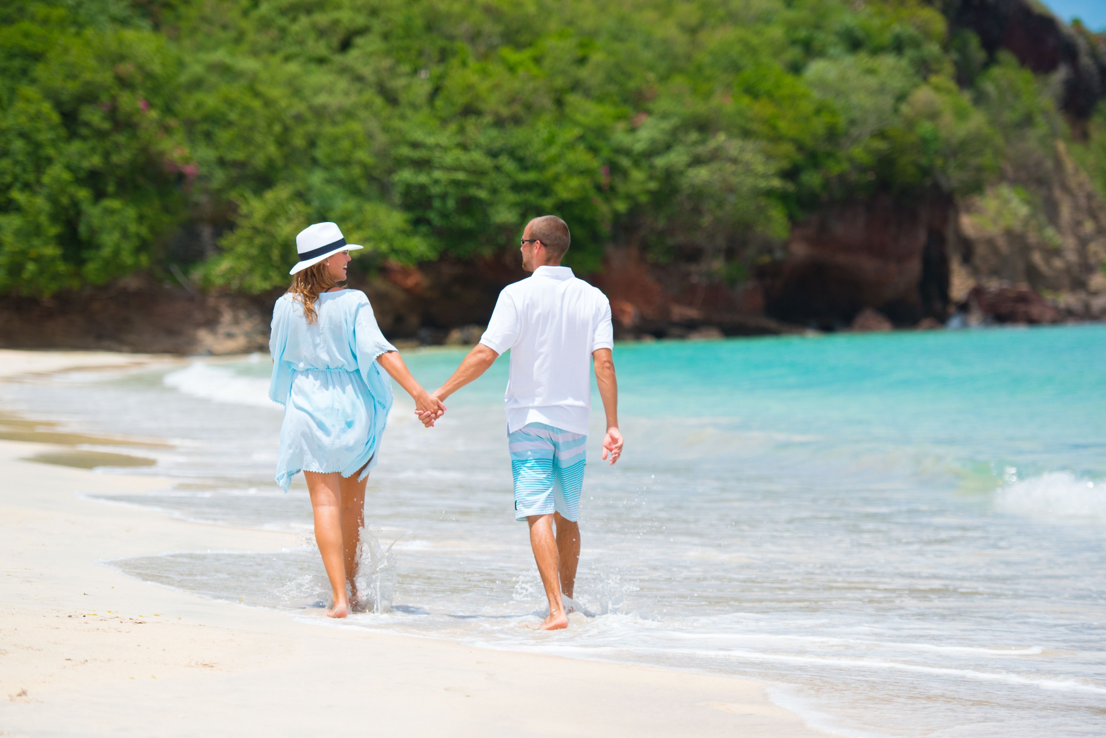 A couple holding hands walking on a beach with clear turquoise water and greenery in the background.