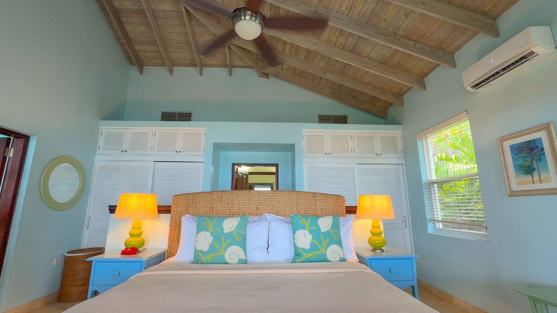 A bedroom with a wooden ceiling, two white louvered closets, a bed with colorful pillows, two bedside lamps, and an air conditioning unit.