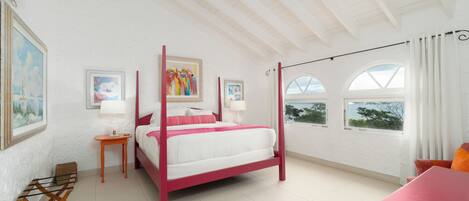 A spacious bedroom with a four-poster bed, a sitting area with a table and chairs, and large windows offering a view of the sea.