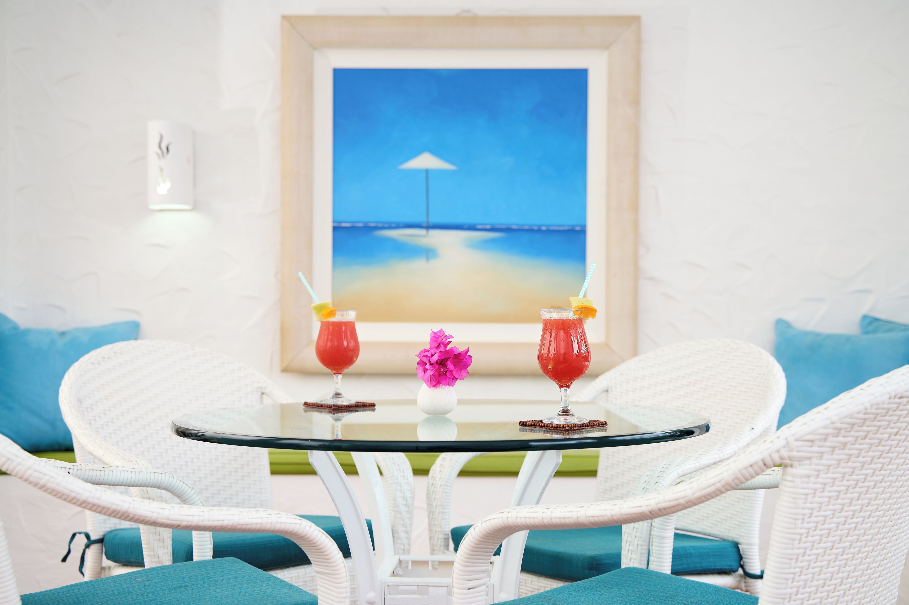 A round glass table with two drinks and a vase of flowers, surrounded by white wicker chairs with teal cushions, set against a beach painting.