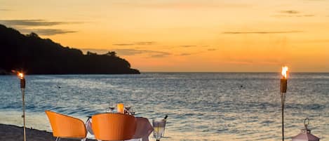 A beachside table with chairs and a candle, set against a sunset view.