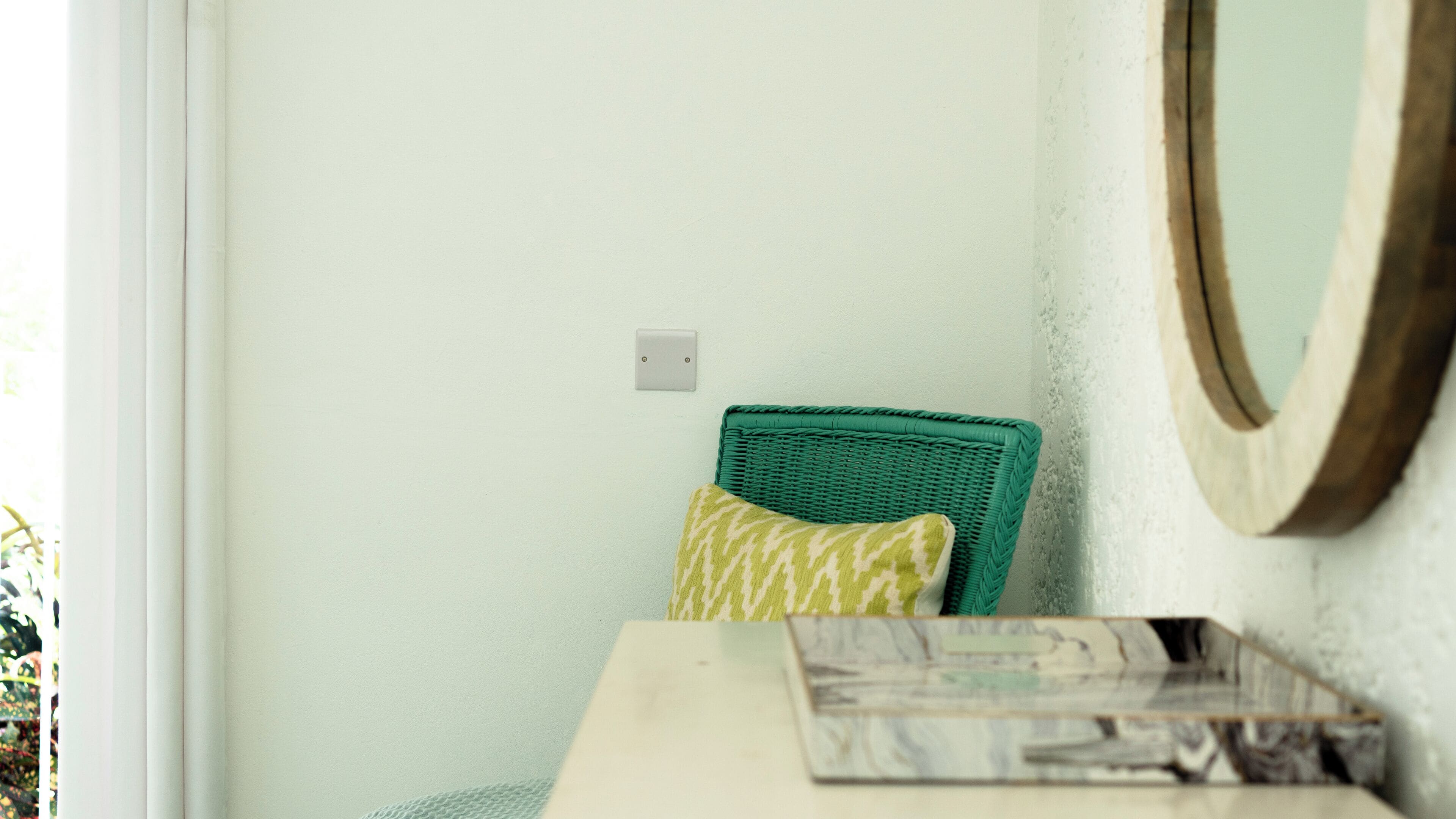 Un rincón de una habitación con una silla de mimbre, un cojín, una mesita con una revista y un interruptor de luz en la pared.
