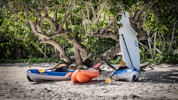 Dos kayaks y una tabla de surf sobre una playa arenosa con follaje denso al fondo.