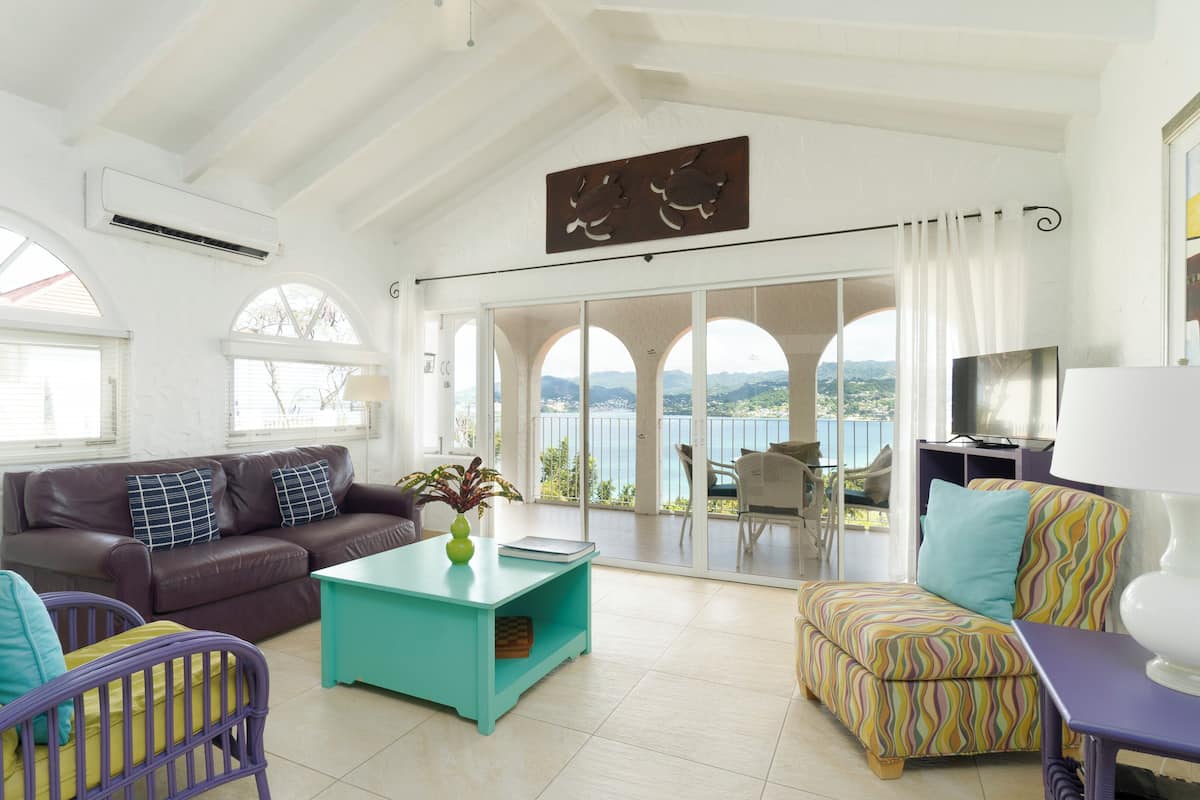A spacious living room with a large window overlooking a scenic view, a comfortable sofa, a teal coffee table, and a colorful ottoman.