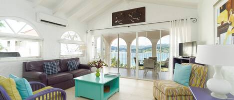 A spacious living room with a large window overlooking a scenic view, a comfortable sofa, a teal coffee table, and a colorful ottoman.