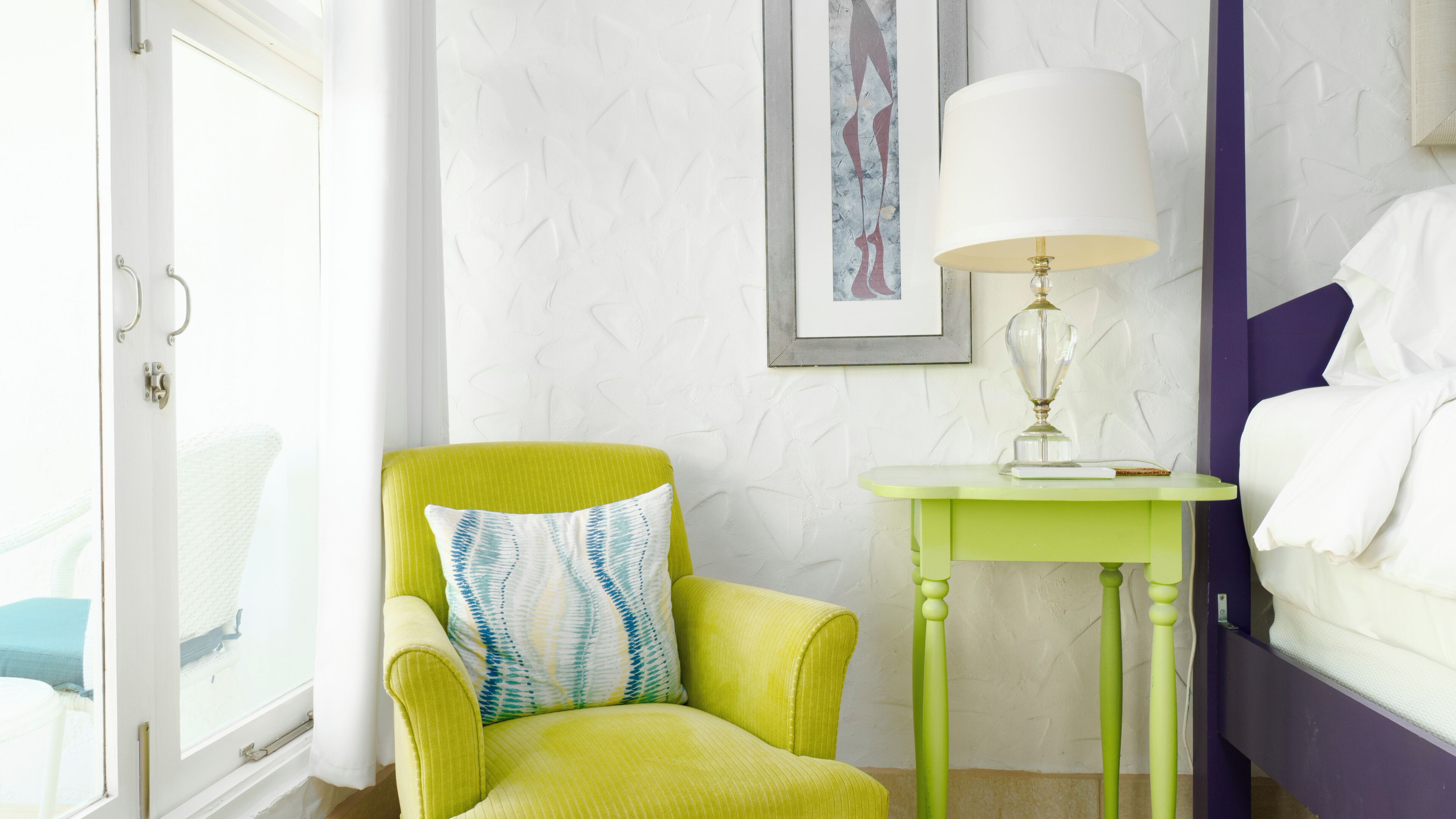 Une pièce avec un fauteuil vert, une petite table avec une lampe, un lit et une fenêtre avec des rideaux.
