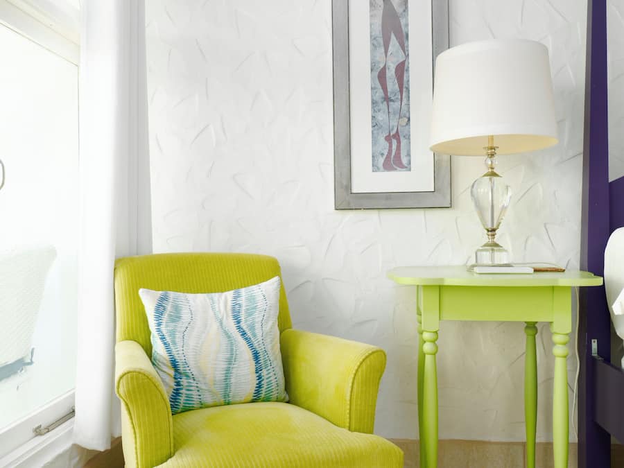 A room with a green armchair, a small table with a lamp, a bed, and a window with curtains.