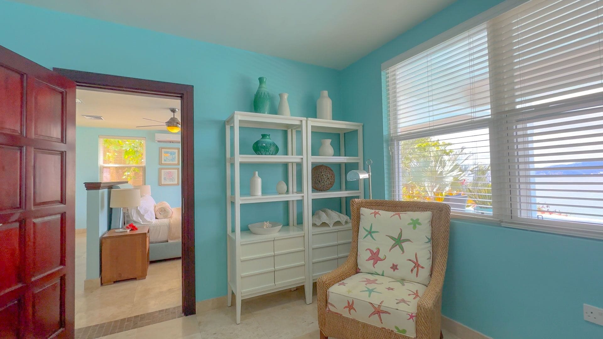 A room with teal walls, a white shelving unit, a wicker chair, and a view through a window with blinds.