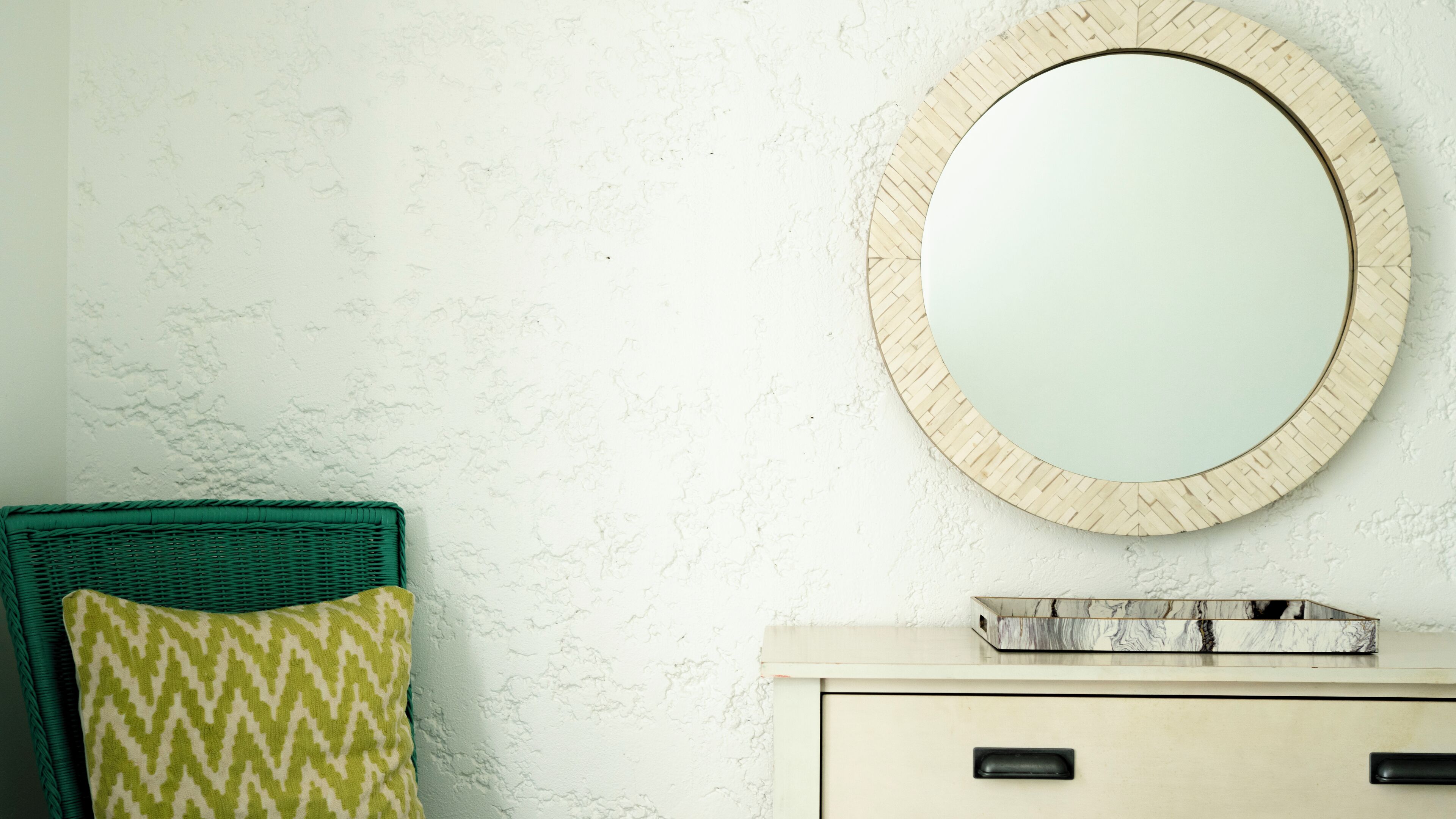 Un baño con un espejo redondo, una silla de mimbre verde y un aparador blanco.