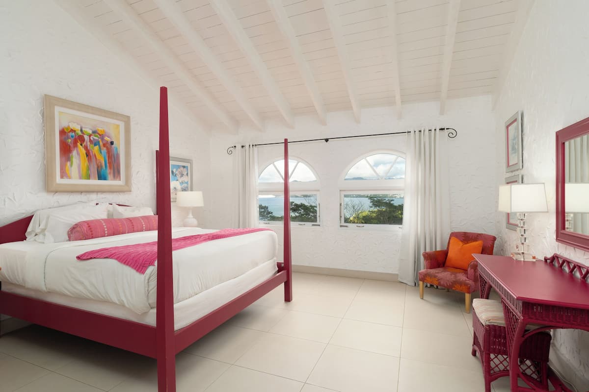A four-poster bed with a white and pink color scheme, a desk with a red chair, and a view of the sea through arched windows.