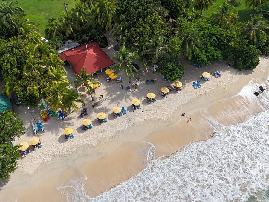 Vista aérea de una playa con filas de sombrillas y sillas de playa, un edificio de techo rojo y una orilla arenosa con olas.