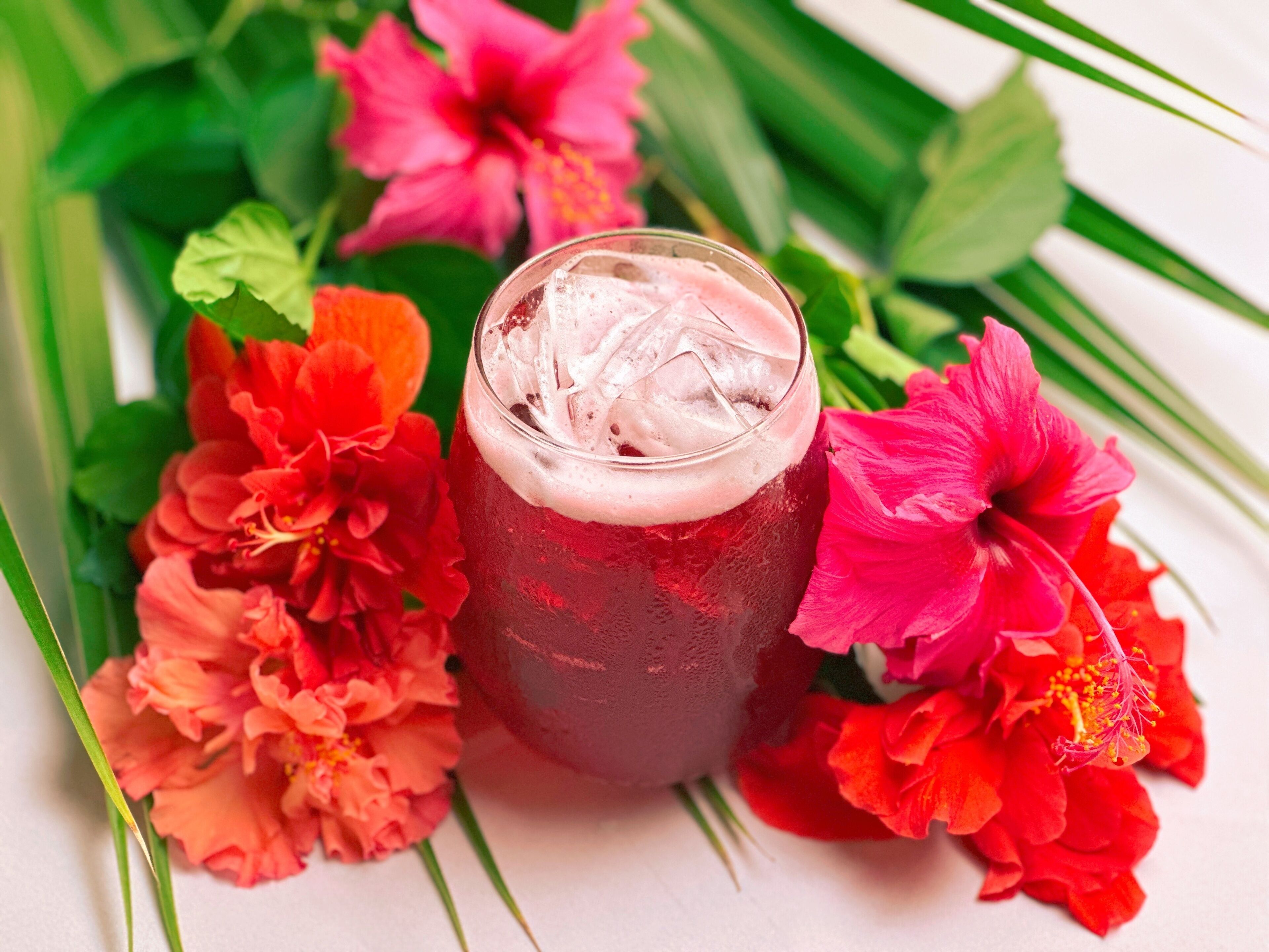 Un bicchiere di bevanda rossa con ghiaccio, circondato da fiori colorati.