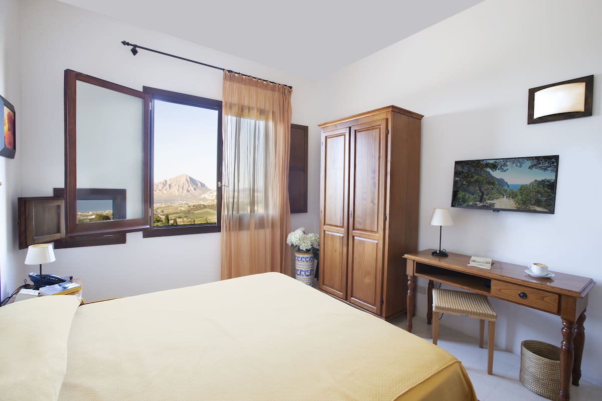 A hotel room with a bed, a desk, a chair, a wardrobe, a window with a view, a lamp, and a vase with flowers.
