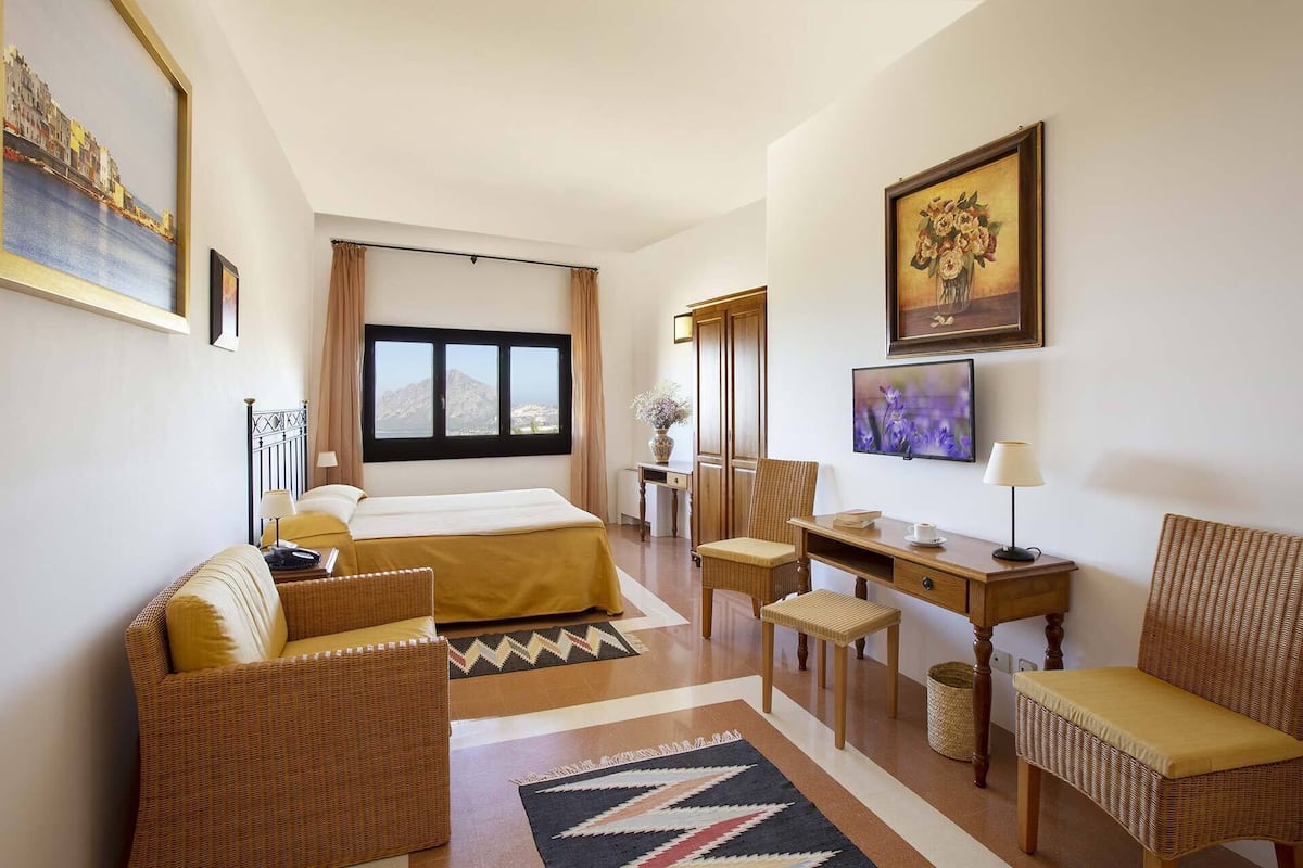 A hotel room with a bed, a desk, two chairs, a TV, and a window with a mountain view.
