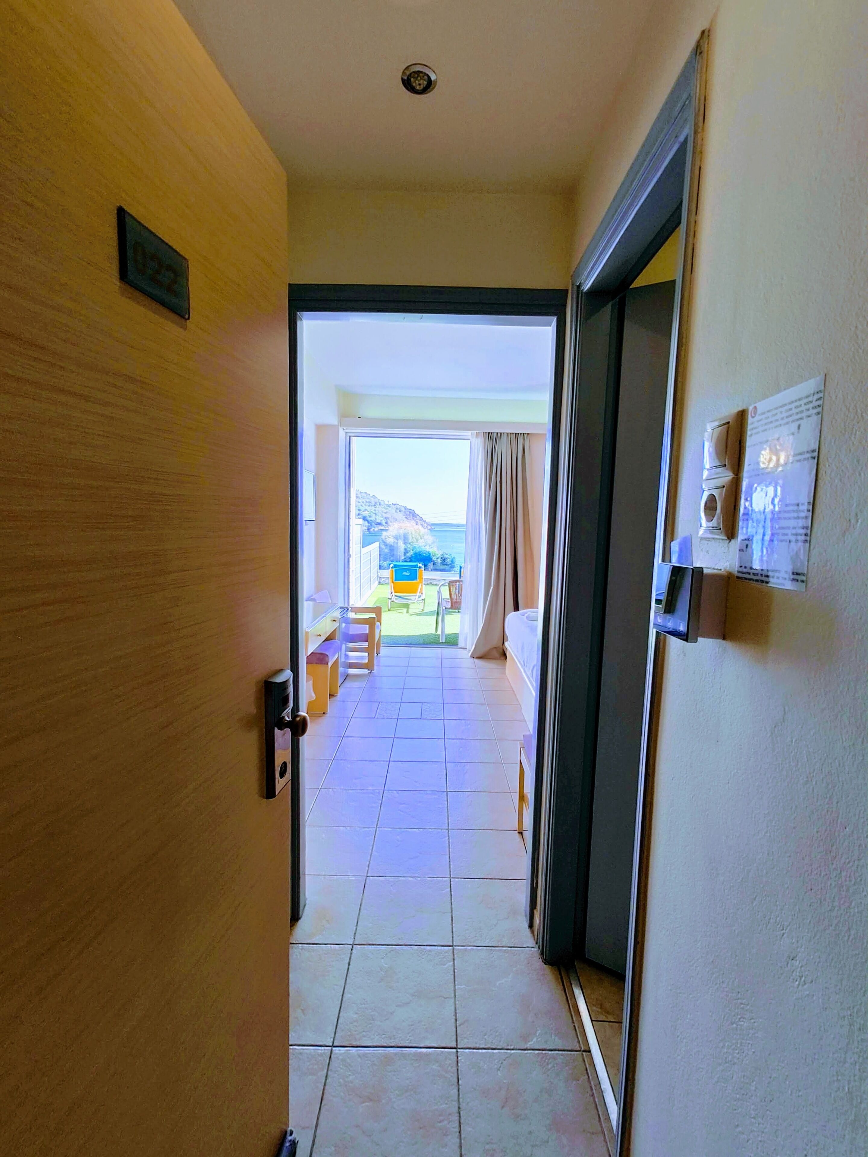 Habitación de hotel con puerta de madera, vista a un balcón con mesa y sillas, y piso de baldosas.