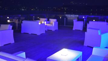 A rooftop lounge area with white wicker furniture and a small table with a light.
