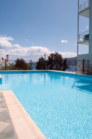 A clear blue swimming pool with a tiled edge, surrounded by a fence and a building with balconies. - New Aegli Resort Hotel (Poros)