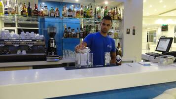 Zona de bar con un barman preparando bebidas, una máquina de café y una variedad de botellas de licor.