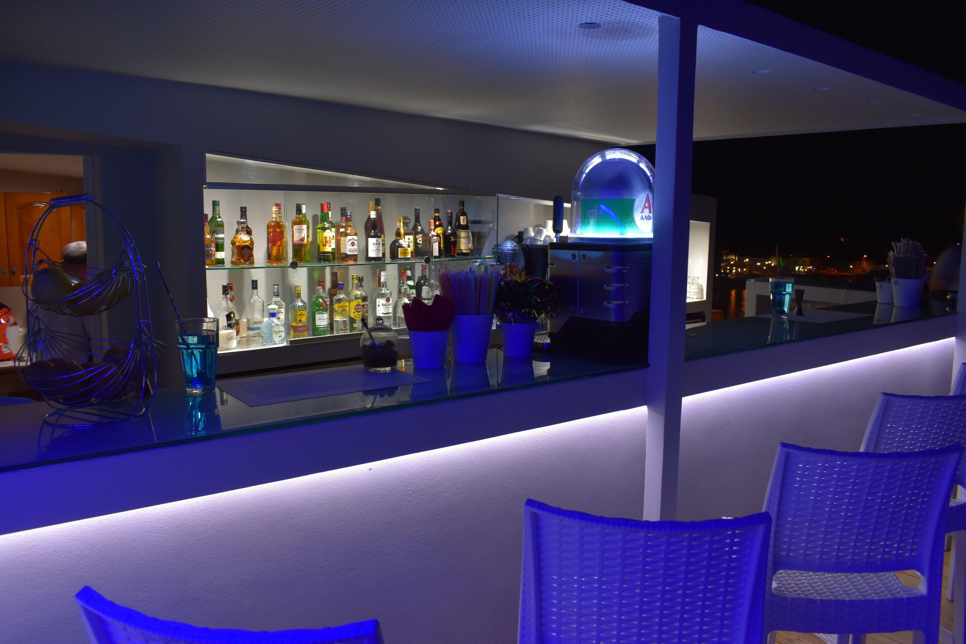Un espace bar doté d’un comptoir en verre, d’étagères remplies de bouteilles d’alcool et d’une enseigne au néon.