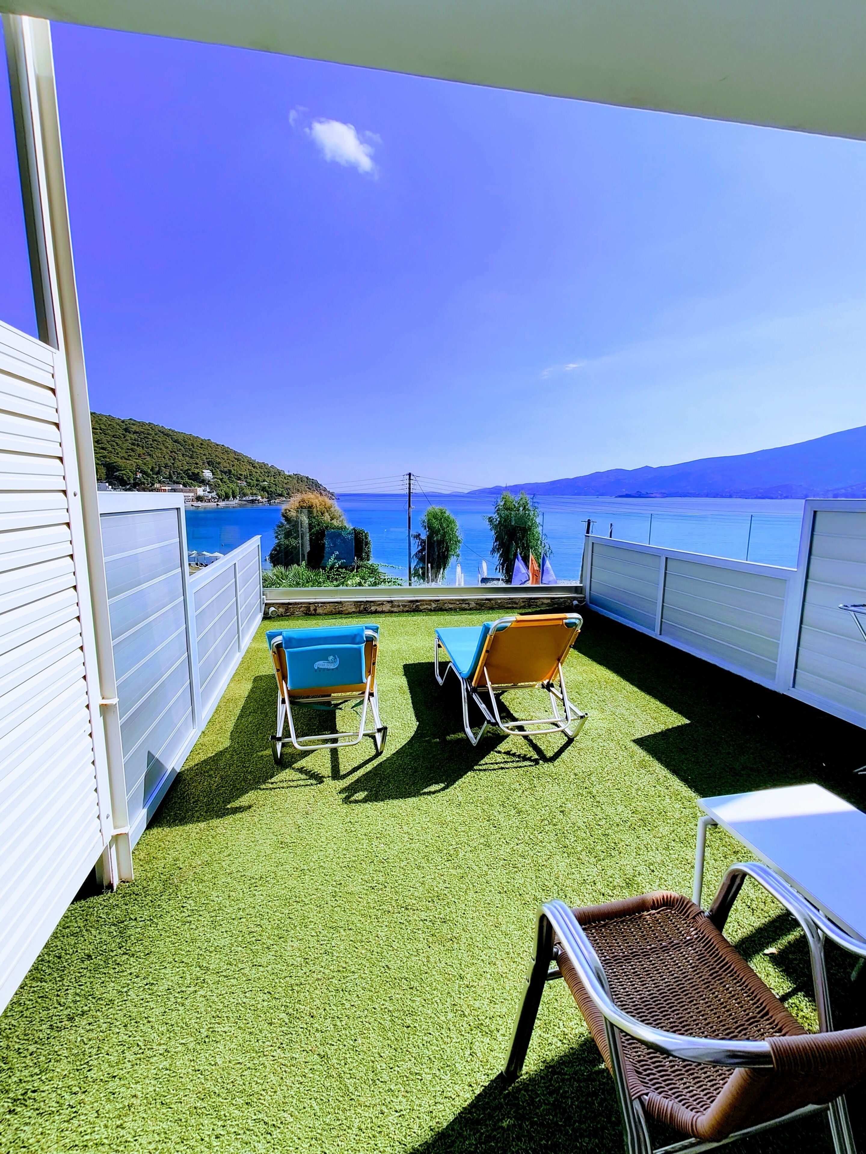 A balcony with artificial grass, two blue chairs, a wicker chair, and a table.