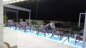 Een terras met rieten stoelen en een glazen tafel, verlicht met inbouwverlichting.
