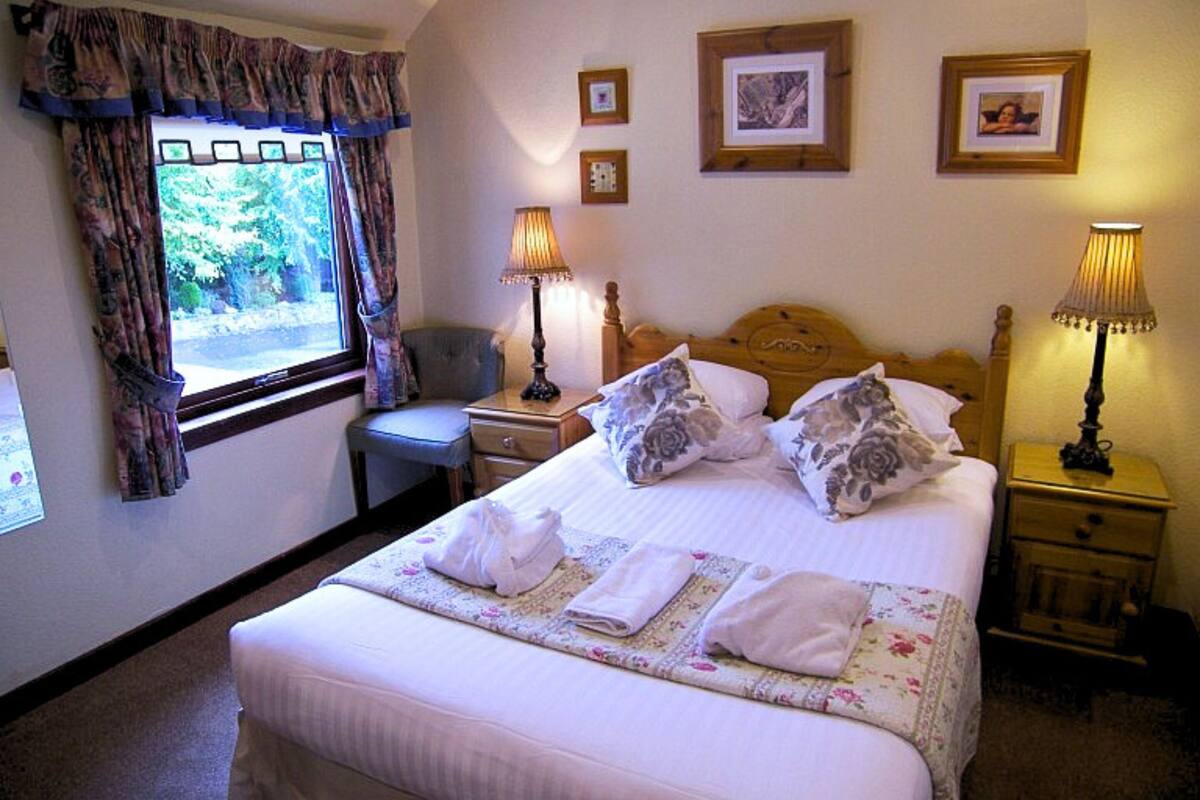 A neatly made bed with pillows and folded towels, a bedside table with a lamp, a chair, and a window with curtains.