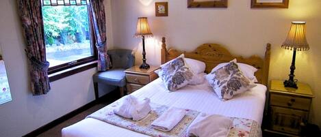 Una cama bien hecha con almohadas y toallas dobladas, una mesita de noche con lámpara, una silla y una ventana con cortinas.