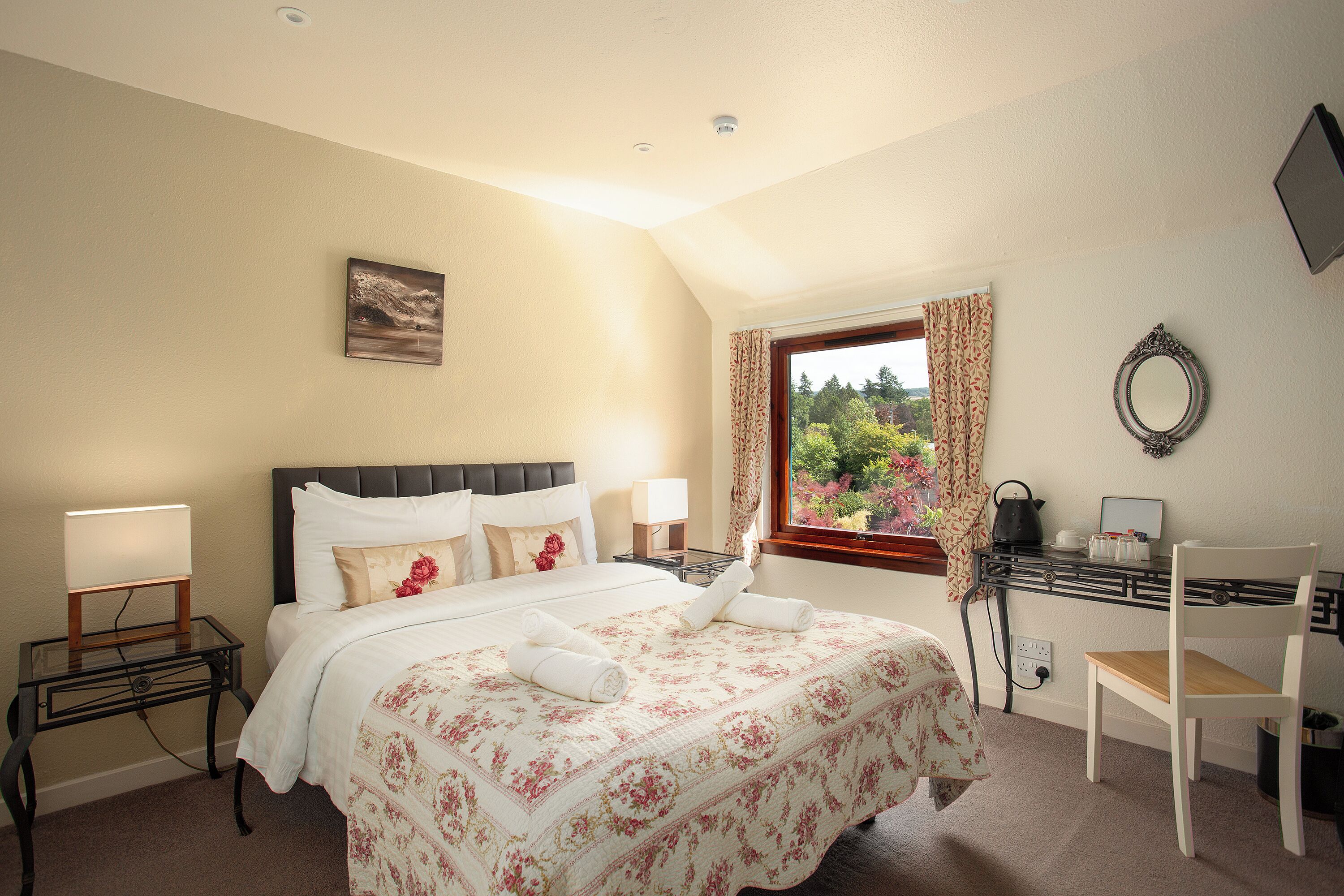 A bedroom with a bed, bedside tables, a desk with a chair, a window with curtains, and a view of greenery outside.