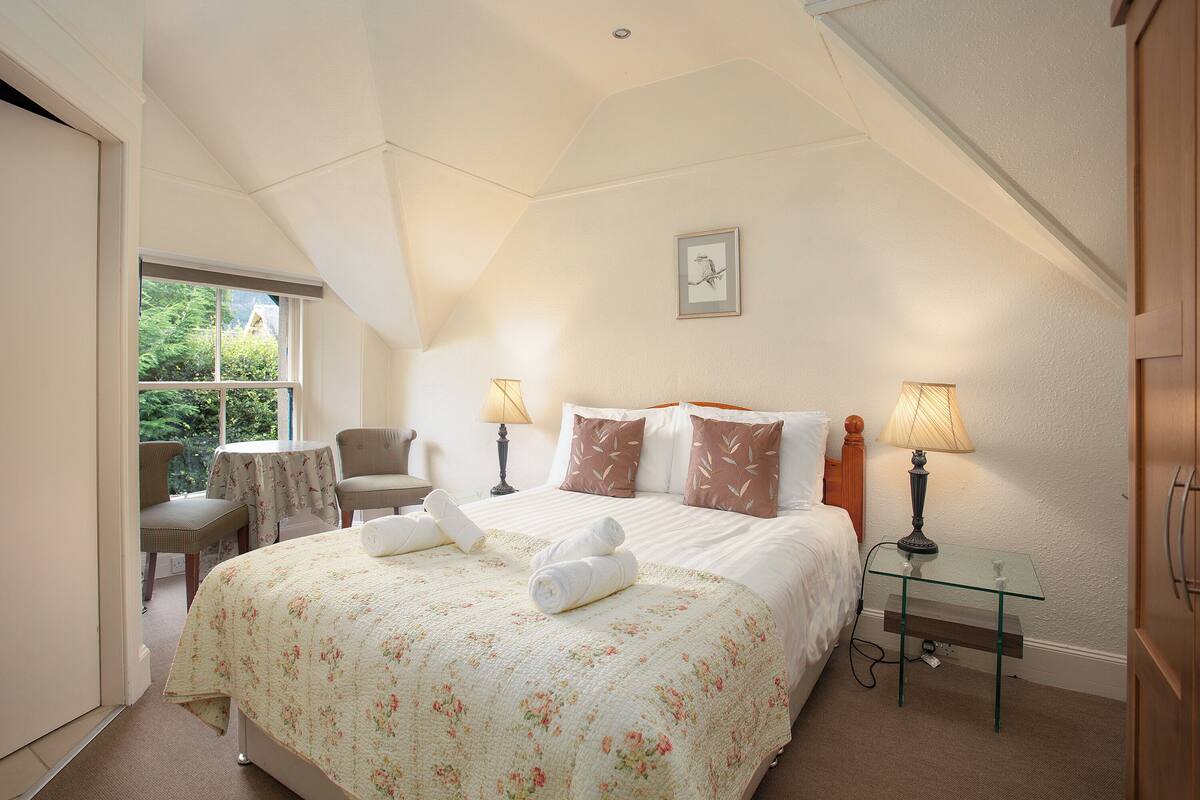 A bedroom with a large bed, two bedside lamps, a small table, a chair, and a window with a view of greenery.