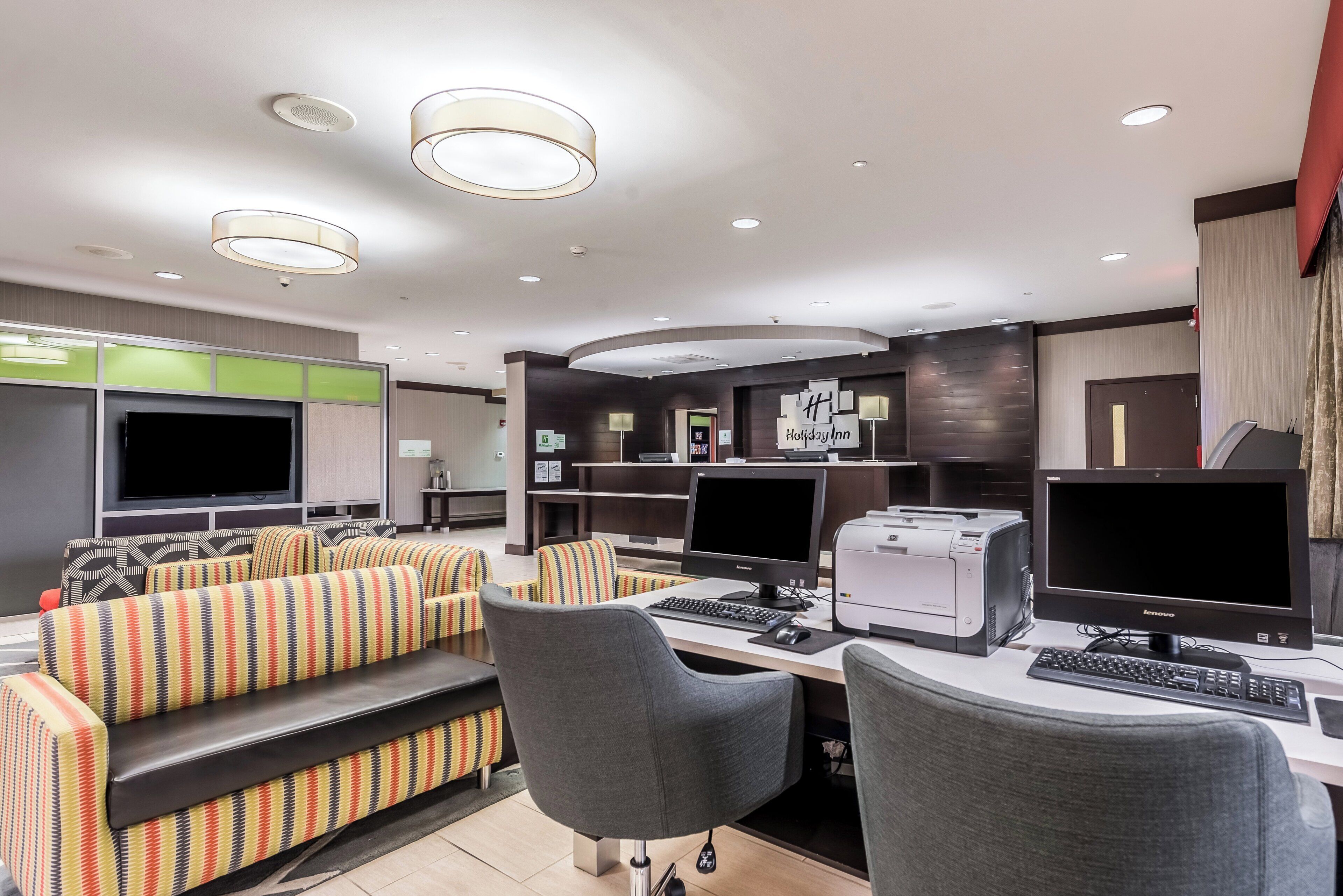 Een moderne hotelkamer met een bank, twee werkplekken met computers, een flatscreen televisie en een bureau met een printer.