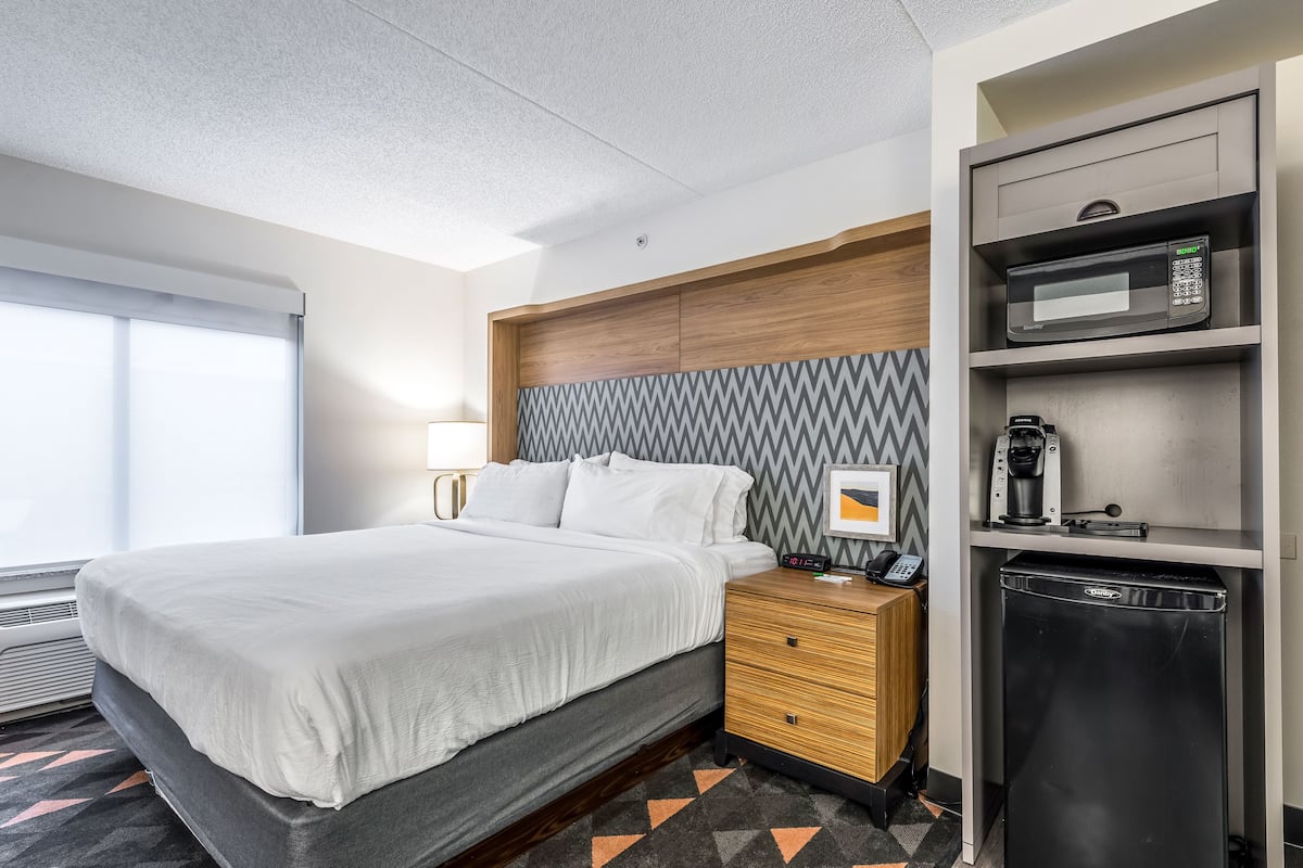 A hotel room with a bed, bedside table, microwave, coffee maker, and a small refrigerator.
