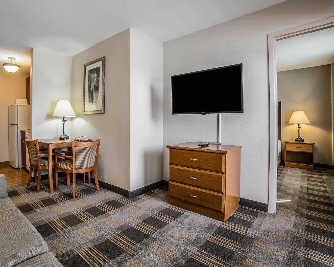 A hotel room with a flat-screen TV, a wooden dresser, a dining table with chairs, and a patterned carpet.