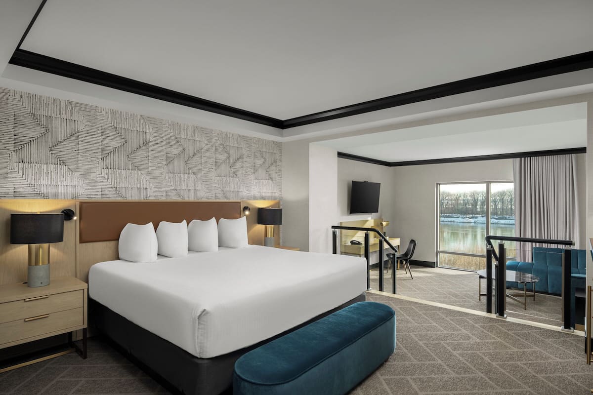 Habitación de hotel con una cama grande, un escritorio con silla, televisor y balcón con vista al lago.