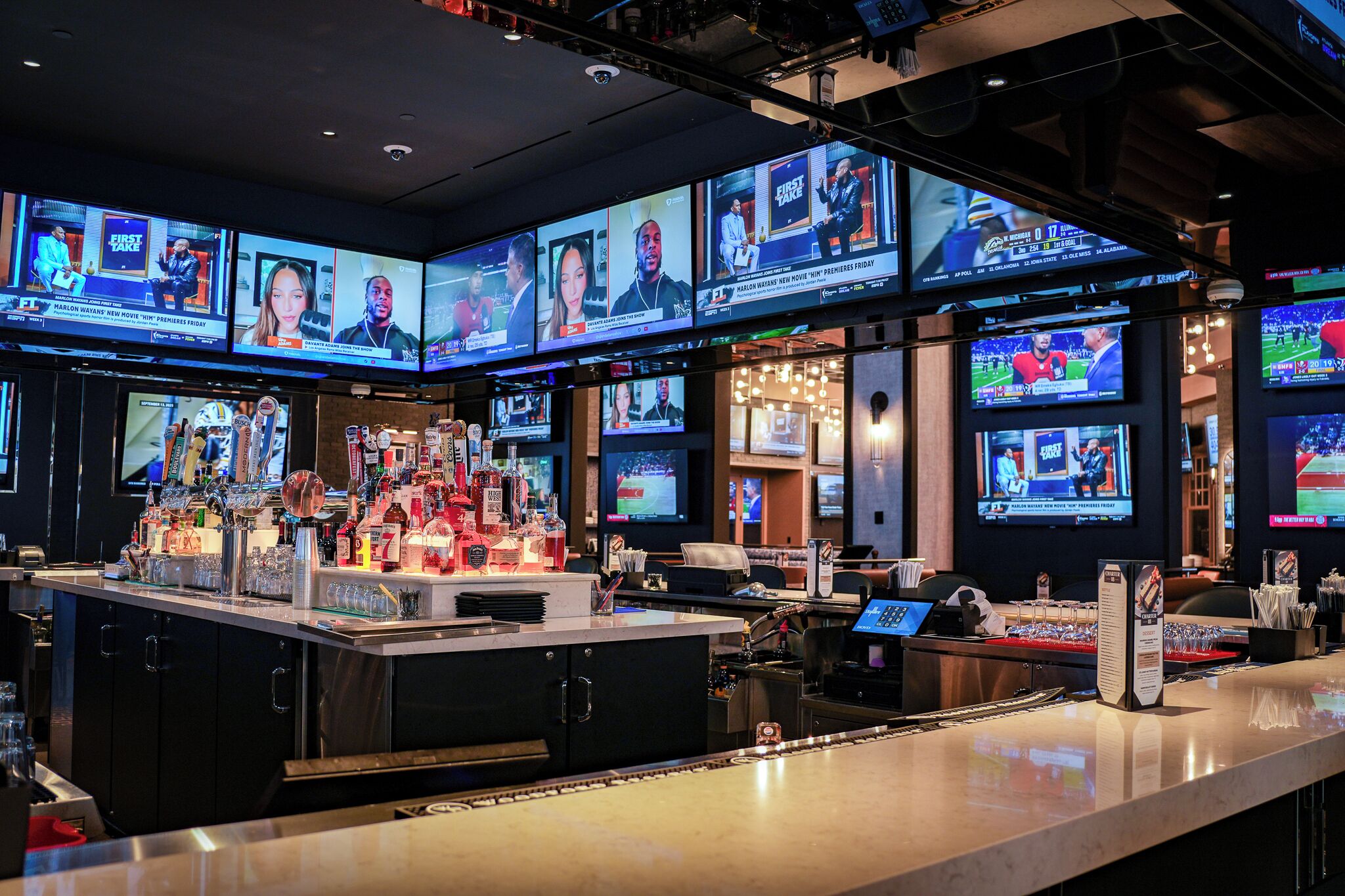A bar area with multiple screens displaying various content, a well-stocked bar counter, and a seating area with tables.