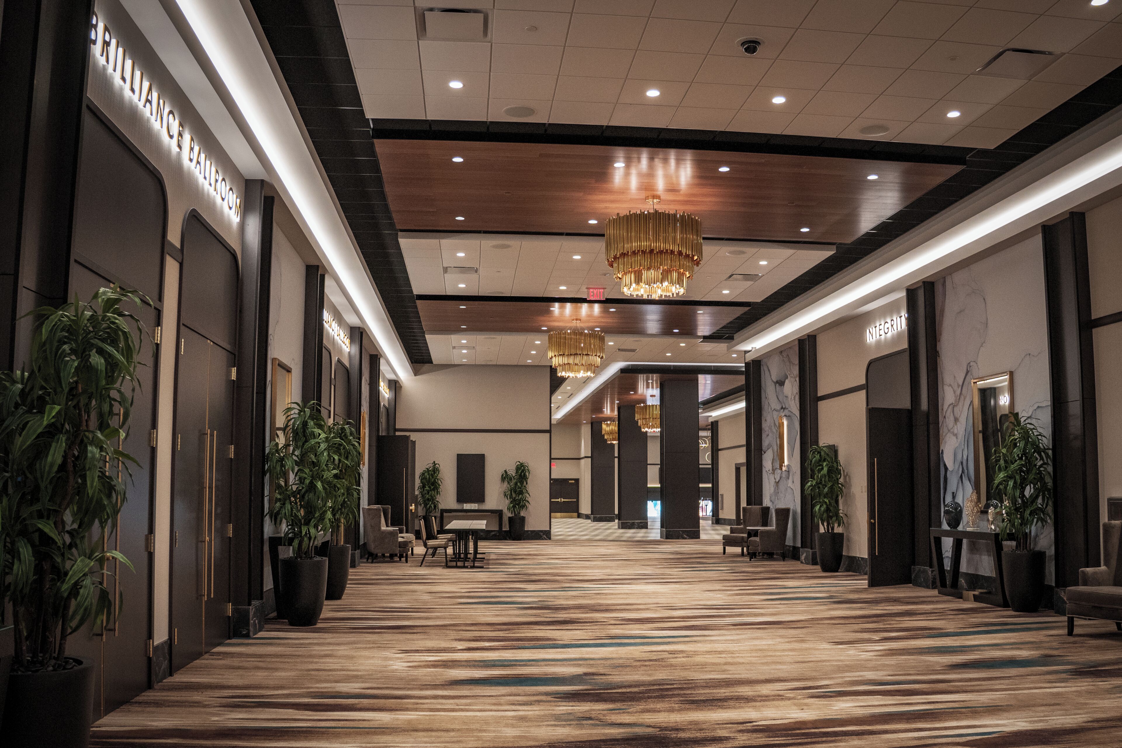 A spacious hotel lobby with a long corridor, large potted plants, chandeliers, and a marble wall with the word 'INTEGRITY'.